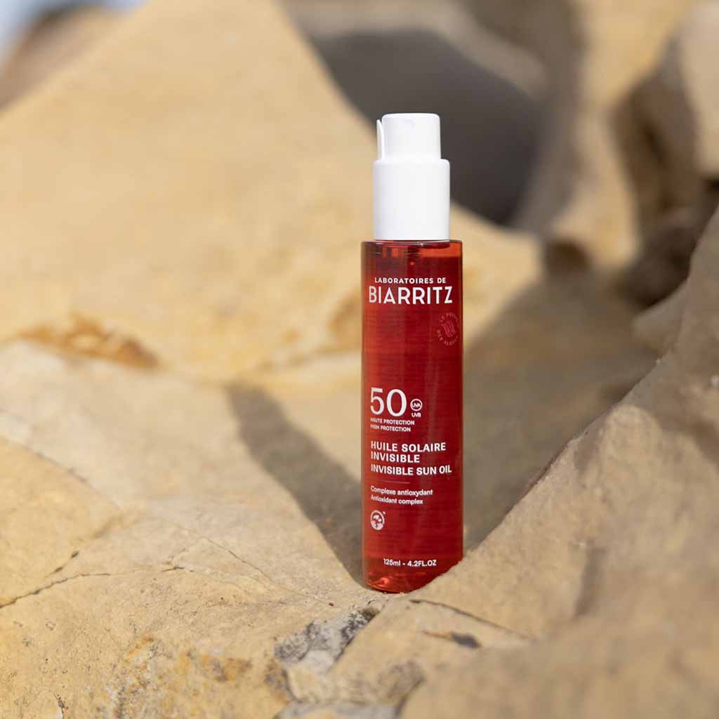 Laboratoires de Biarritz Suncare Invisible Oil SPF50; pullo Biarritz Invisible Sun Oil SPF 50 -öljyä seisoo pystyssä beigellä kivellä auringonvalon valaisemana ja epätarkkaa kivistä taustaa vasten; pullo sylinterin muotoinen puoliläpinäkyvää tai huurteista muovia kiiltopinnalla, oranssi pumppukorkki; valkoinen groteskikirjasin näyttää brändi- ja tuotenimen etuetiketissä; ei ihmisiä tai näkyvää toimintaa kuvassa.