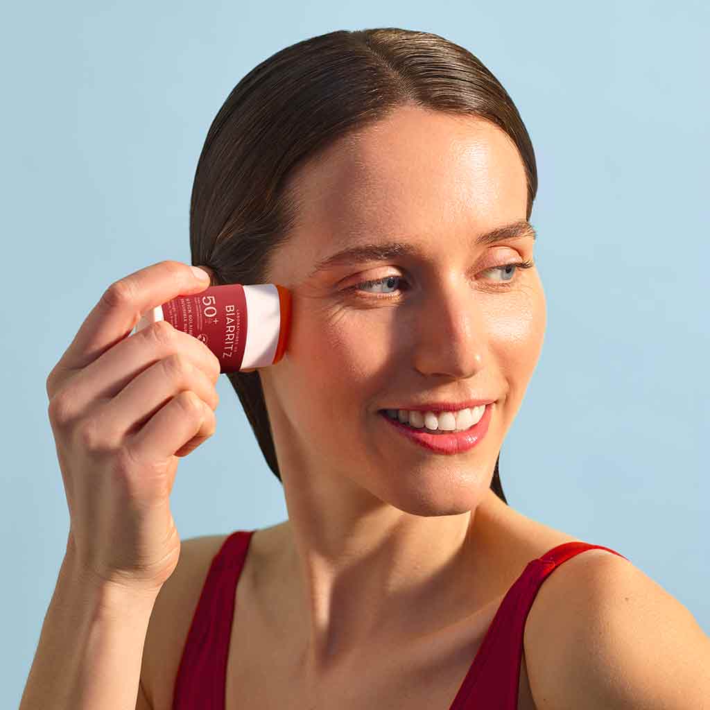 Laboratoires de Biarritz Suncare Invisible Sun Stick SPF50+; henkilö punaisessa yläosassa hymyilee levittäessään aurinkosuojapuikkoa kasvoilleen vaaleansinistä taustaa vasten; aurinkosuoja sylinterin muotoisena kiinteänä puikkona muovipakkauksessa, mattavalkoinen korkki ja värillinen pohja; yksinkertaista groteskitekstiä näkyvissä etiketissä; ei muita esineitä tai rekvisiittaa; kuva vyötäröstä ylöspäin, tarkennus levitystoiminnossa.
