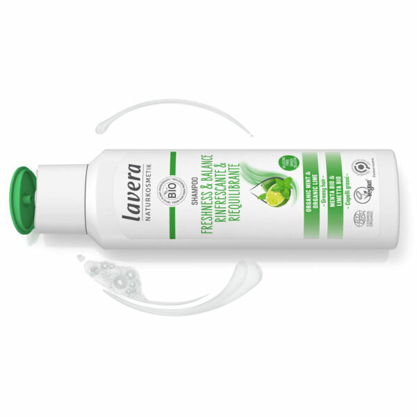 Valkoinen Lavera-shampoopullo vihreällä läppäkorkilla, nimellä Freshness & Balance, sisältää luomulimeä ja minttua, viereen aseteltuna raikkaita minttuisia shampoopyörteitä; pullo on muovia sileäpintaisena, pystysuora lieriömäinen muoto; etiketissä groteskikirjasin eri painoin; tuote kuvattu tasaisella taustalla ilman ihmisiä tai rekvisiittaa.