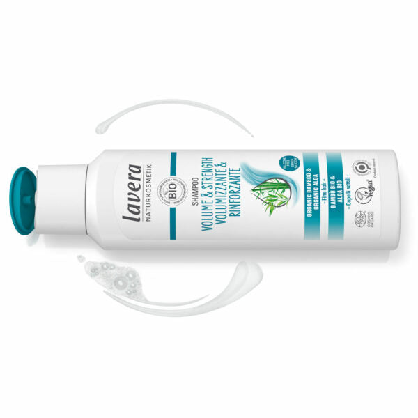 Valkoinen Lavera Volume & Strength Shampoo -pullo minttuetiketillä lepää valkoisella alustalla, korkki on turkoosi ja avoinna; pakkaus kiiltäväpintaista muovia, lieriömäinen pyöristetyllä olkaosalla ja tasaisella pohjalla; ympärillä näkyy shampoolammikoita ja vaahtopyörteitä; musta ja turkoosi groteskiteksti lukee Volume & Strength Shampoo; kuvassa ei ole ihmisiä tai käsiä.