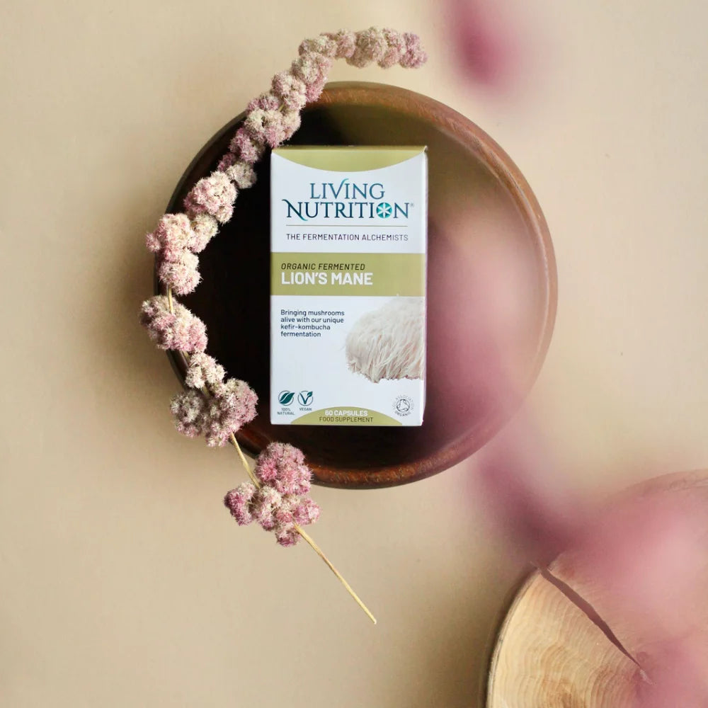 Living Nutrition Fermentoitu Lions Mane; suorakulmainen laatikko merkitty "Living Nutrition Organic Fermented Lions Mane" seisoo pystyssä luonnonvaalean ruskean puisen kulhon sisällä; useita kuivattuja vaaleanpunaisia kukkia on aseteltu laatikon ympärille kulhossa; alusta on beige ja tasainen mattapinnalla; kuva otettu ylhäältä ilman ihmisiä tai käsiä.