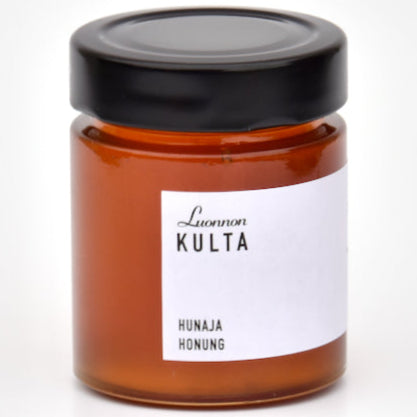 Luonnon Kulta Juokseva Hunaja; pieni lasipurkki täynnä kultaista hunajaa, mustalla kannella ja valkoisella etiketillä, jossa lukee Luonnon Kulta ja Hunaja Honung; kirkas lasipurkki on sylinterinmuotoinen sileällä, kiiltävällä pinnalla; musta kansi on litteä ilman näkyviä merkintöjä; valkoisessa paperietiketissä lihavaa groteskitekstiä tummalla värillä; tausta valkoinen, kuvattu studiovalaistuksessa; ei ihmisiä tai käsiä näkyvissä.