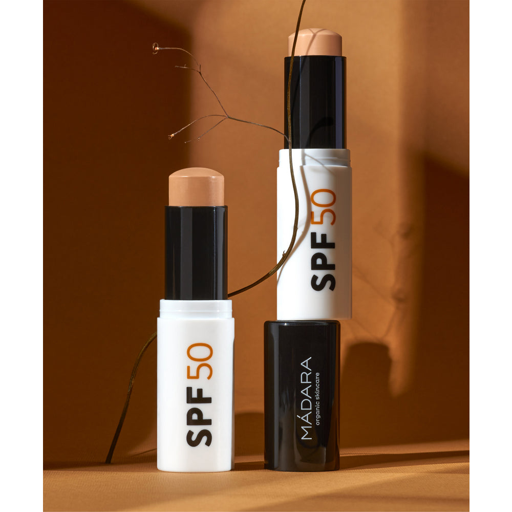 Mádara Mineral SPF50 Sunscreen Stick; kaksi SPF 50 -aurinkosuojapuikkoa ja suljettu MÁDARA-puikko on aseteltu ruskealle pinnalle kuivan kasvinvarren viereen, lämpimän ruskeaa taustaa vasten; aurinkosuojapuikot ovat sylinterinmuotoisia mattabeigen pakkauksen kera, toisessa lukee "SPF 50"; MÁDARA-puikko on sylinterinmuotoinen mattavihreä; yksinkertainen groteskiteksti valkoisena ja tummana; puikkojen korkit kiinni; ei käsiä tai ihmisiä kuvassa.
