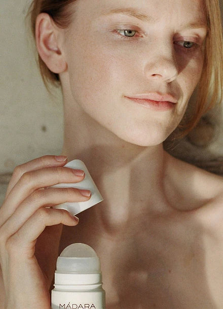 Madara Herbal Deodorant; vaaleaihoinen henkilö pitelee avattua MÁDARA-roll-on-tuotetta kasvojensa lähellä katsoen sivulle mietteliäänä; lieriömäinen tuote valkoisella mattapinnalla ja pyöristetyllä levittimellä; musta versaali groteskityyppografia ”MÁDARA” selkeästi pakkauksessa; tausta tasainen, neutraali ja pehmeästi valaistu.