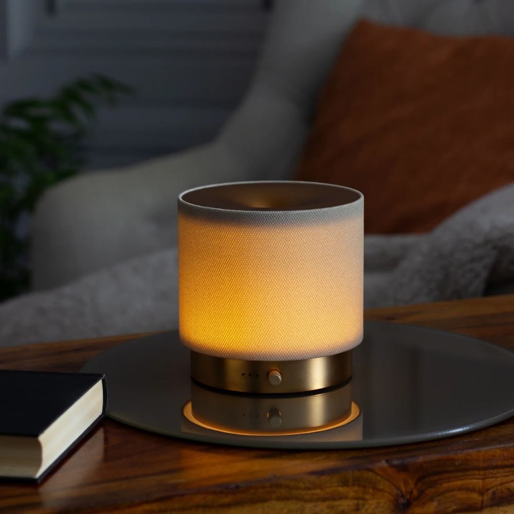 Made by Zen Mira USB Diffuuseri; lieriömäinen lamppu pehmeine lämpimine hehkuineen seisoo pyöreällä peilillä puupinnan päällä; lampun runko läpinäkymätön ilman näkyvää brändäystä tai merkintöjä; pinta mattapintaista puunsyistä, peili pyöreä ja heijastavapintainen; suljettu muistivihko kiinteine kansineen lampun vieressä; taustalla epätarkka neutraalinvärinen kangassohva ja oranssi tyyny; ei ihmisiä tai käsiä kuvassa.