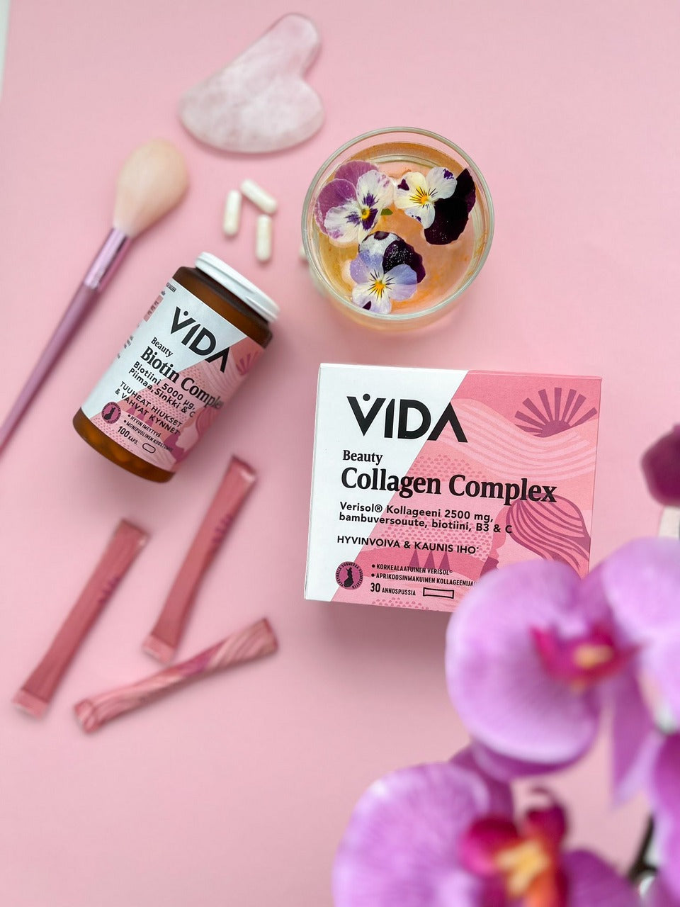 Vida Beauty Biotin Complex; lieriön muotoinen pullo ja suorakulmainen laatikko tekstillä "VIDA Collagen Complex" sans-serif-fontilla, useita yksittäin käärittyjä vaaleanpunaisia ravintolisätikkuja, valkoisia kapseleita, metallinvärinen vaaleanpunainen meikkisivellin pehmein harjaksin, kirkas lasimaljakko pienin kukin ja vaaleanpunainen orkidea aseteltuna pastellinvaaleanpunaiselle mattapinnalle; ei ihmisiä tai käsiä.