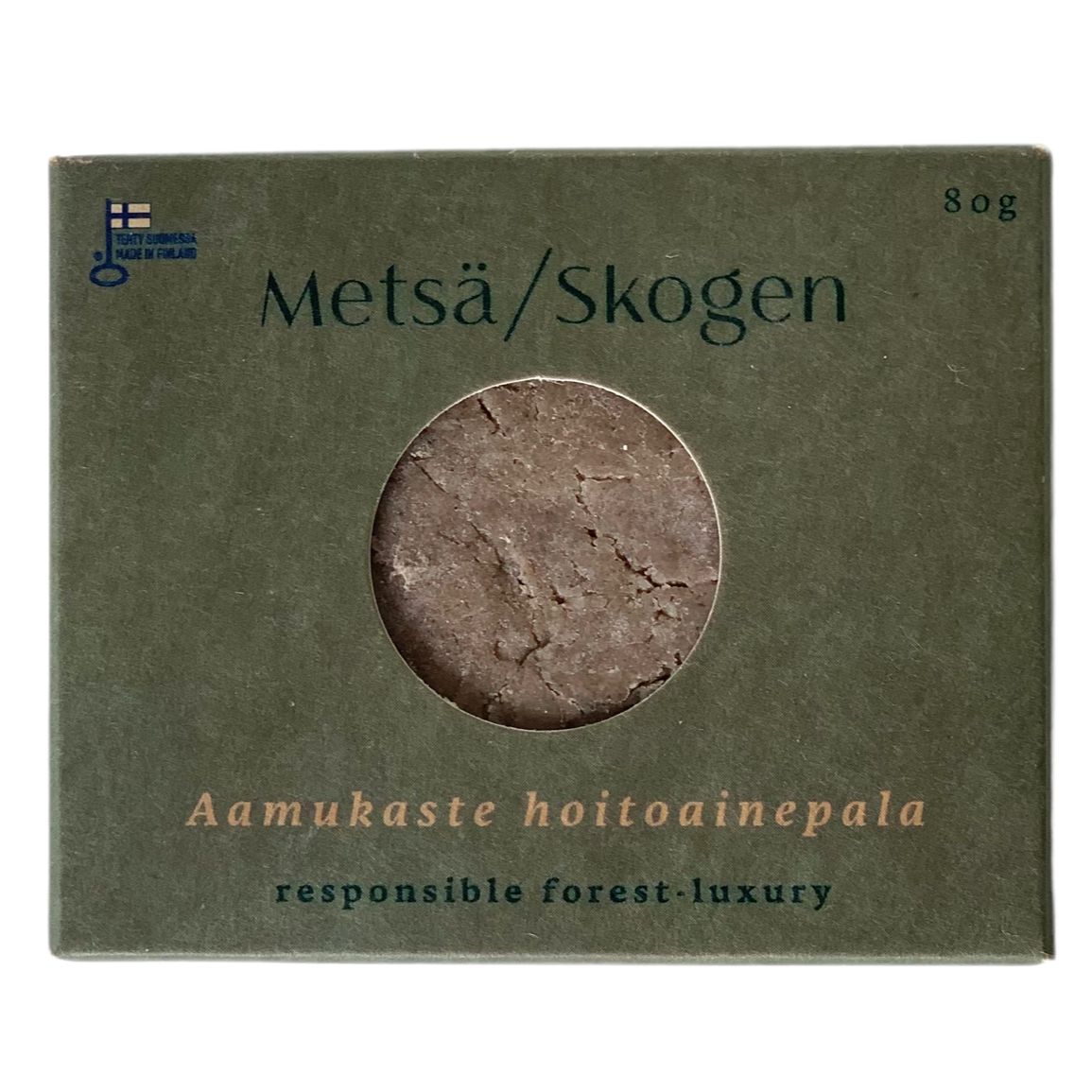 A green box with a round window displays a brown solid bar. The box is rectangular with a matte finish and has white, sans-serif text reading Metsä/Skogen, Aamukaste hoitoainepala, and responsible forest-luxury. No people or hands are present. The background is plain and evenly lit, and the shot is taken straight on at eye level. This packaging reflects an eco-conscious, Scandinavian-inspired aesthetic.