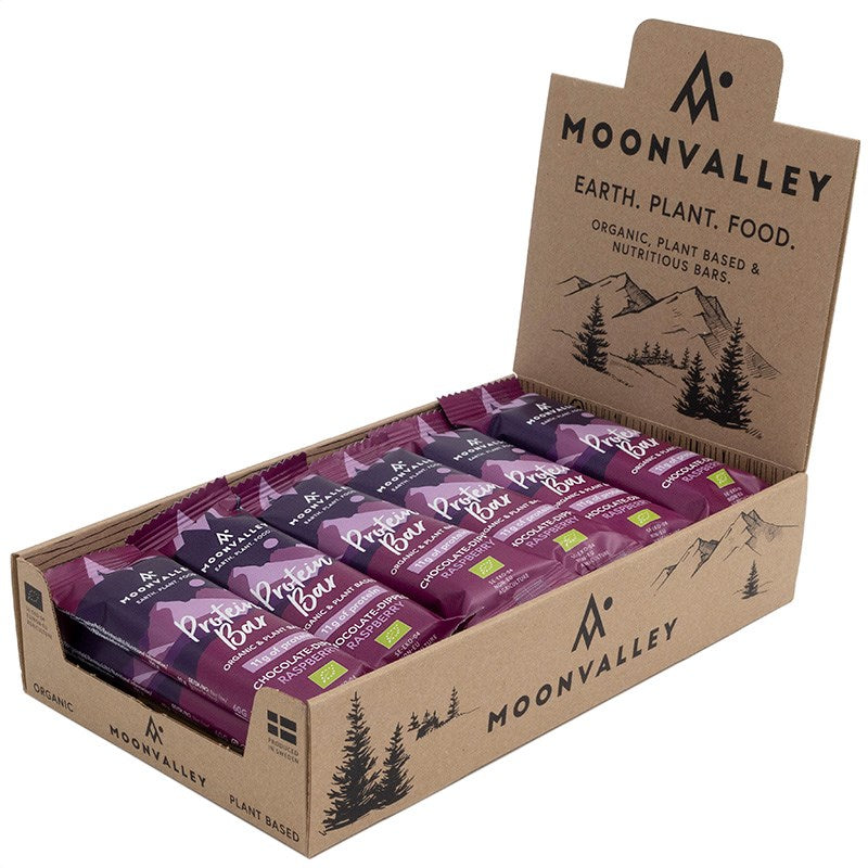 Moonvalley Luomu Proteiinipatukka Vadelma; avattu pahvinen esittelylaatikko Moonvalley-suklaa- ja merisuolaproteiinipatukoita; suorakulmainen laatikko avoimella läpällä mattapinnalla, kannessa vuori- ja luontokuvituksia sekä vasemmalle tasattua lihavoitua groteskitekstiiä ruskean, vihreän ja violetin sävyissä; luettavissa ”organic” ja ”plant-based”; useita violettikääreisiä patukoita pinottuna sisällä vaalealla taustalla, ei ihmisiä.