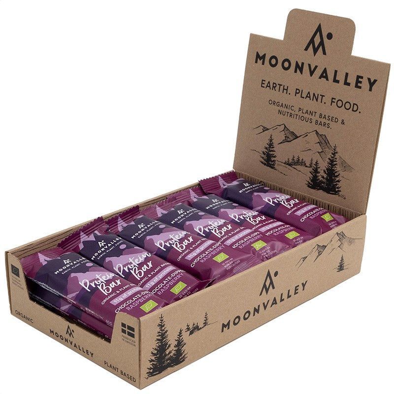 Moonvalley Luomu Proteiinipatukka Vadelma; avattu pahvinen esittelylaatikko Moonvalley-suklaa- ja merisuolaproteiinipatukoita; suorakulmainen laatikko avoimella läpällä mattapinnalla, kannessa vuori- ja luontokuvituksia sekä vasemmalle tasattua lihavoitua groteskitekstiiä ruskean, vihreän ja violetin sävyissä; luettavissa ”organic” ja ”plant-based”; useita violettikääreisiä patukoita pinottuna sisällä vaalealla taustalla, ei ihmisiä.