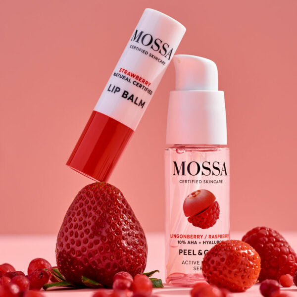 Mossa Berry Kiss Lahjasetti; kaksi ihonhoitotuotetta mansikoin ja vadelmin kuvitettuina; toinen on ravitseva mansikkahuulivoidetuubi, valkoinen ja sylinterinmuotoinen mattapinnalla; toinen on elvyttävä puolukka-vadelmaseerumi pienessä pullossa, vaaleanpunainen etiketti, pumppuannostelija, kiiltäväpinta; molemmissa yksinkertaista groteskibrändäystä; punaiset marjat täydentävät kuvaa pehmeää vaaleanpunaista taustaa vasten.
