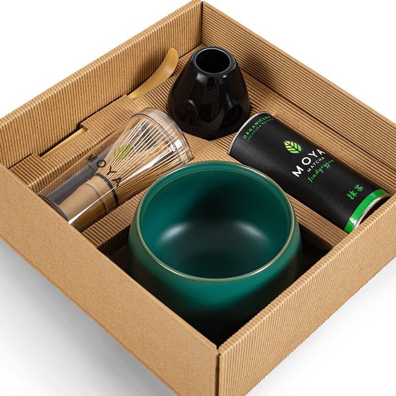 A cardboard gift box contains a bamboo whisk, green tea canister, black whisk holder, bamboo scoop, and a matcha bowl arranged neatly inside. The box is brown with no exterior markings visible. The bamboo whisk has a light wood finish; the black whisk holder is glossy and curved. The green tea canister is metal with simple printed text on the front. All items rest in fitted compartments within the box against plain cardboard backing. No people or hands are shown in the overhead product shot.
