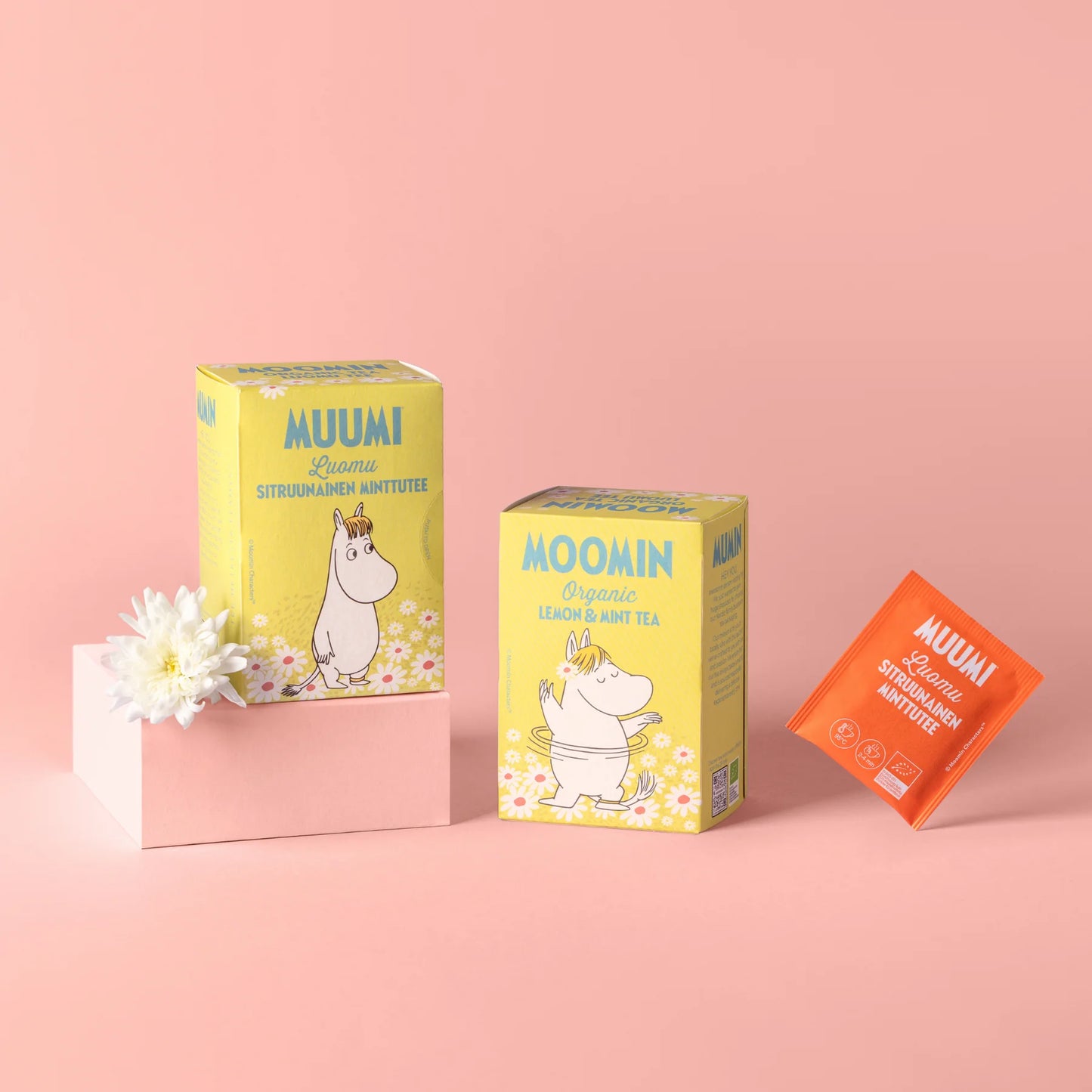 Muumi Luomu Sitruunainen Minttutee: kaksi keltaista Muumin luomusitruuna- ja minttuteerasiaa sekä oranssi teepussi aseteltuna pinkille pinnalle valkoisen kukan kanssa vaaleanpinkkiä taustaa vasten; molemmat rasiat ovat suorakulmaisia mattapakkauksia värikkäin kuvituksin, luettavissa tuotenimi yksinkertaisella pyöristetyllä kirjasimella; virkistävä sitruuna-minttumaku korostuu iloisessa asetelmassa.