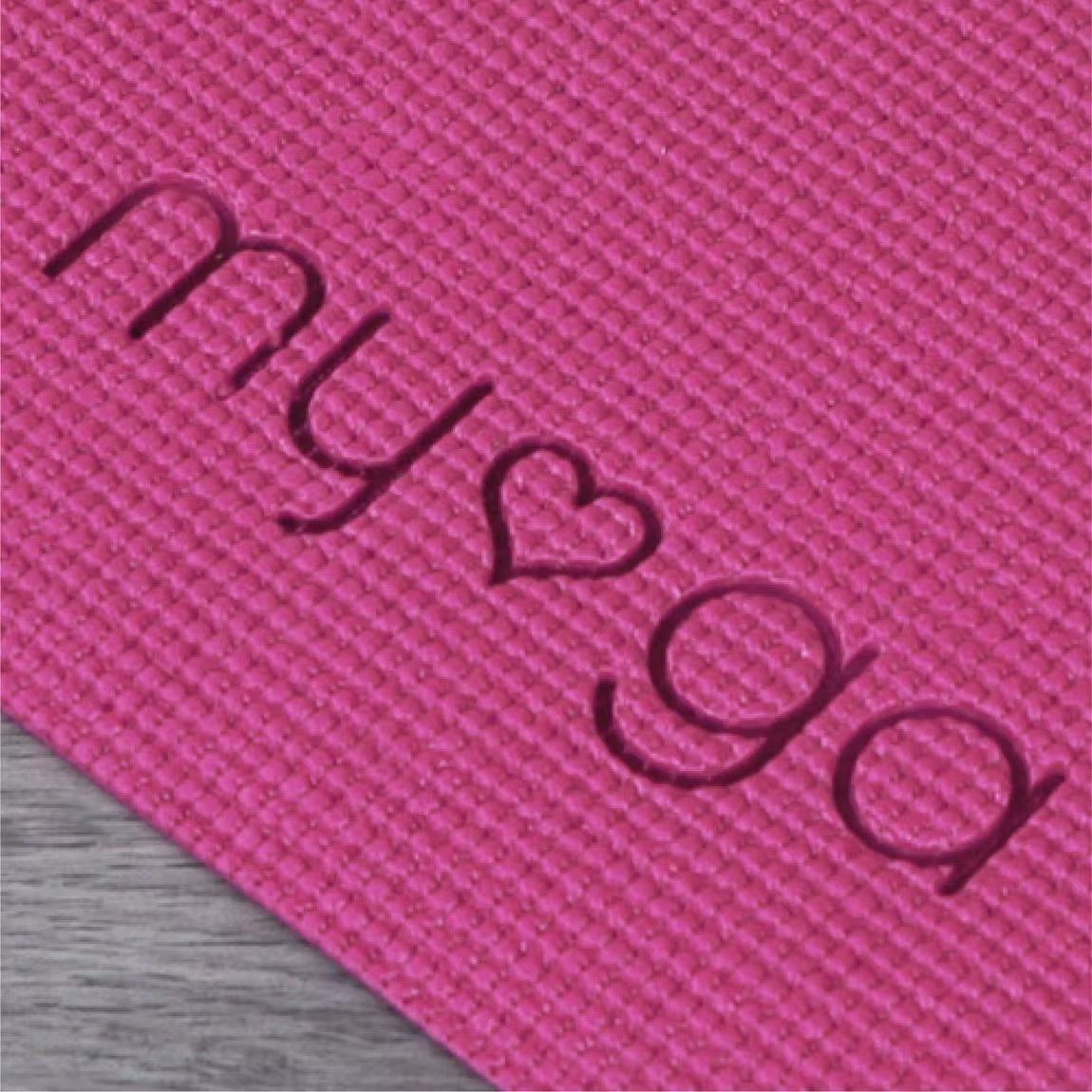 A close-up of a textured pink yoga mat with the word myga embossed, featuring a heart symbol in place of the letter o in yoga, adds a unique and thoughtful detail. The mat has a matte finish and rectangular shape, with no packaging or branding tag visible. The background is neutral and plain. There are no people or hands in the image.