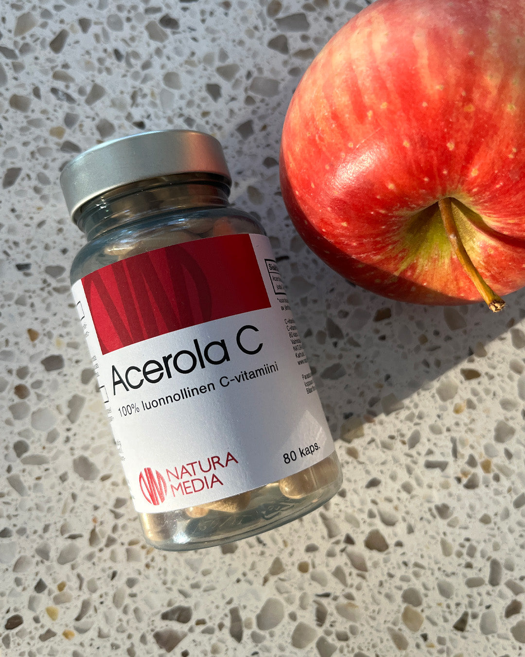 Natura Media Acerola C; valkoinen muovipullo mattapinnalla tekstillä "Acerola C" ja punainen omena lepäävät vaaleasävyisellä, täplikkäällä työtasolla suorassa auringonvalossa; pullo valkoinen korkki ja sylinterin muotoinen; etikettiteksti kertoo "80 capsules, natural vitamin C by Natura Media" korostaen acerolauutteen hyötyjä; ei ihmisiä eikä käsiä näkyvissä; pullo ja omena lepäävät täplikkäällä tasolla auringonvalossa.