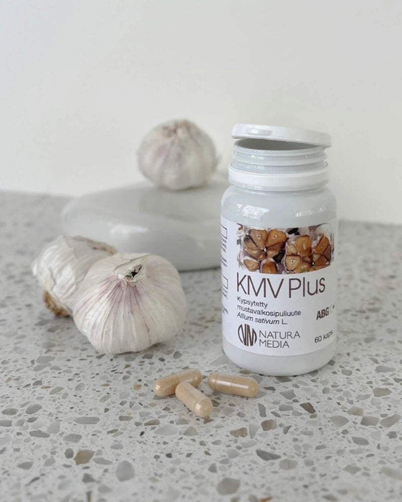 Natura Media KMV Plus; pullo tekstillä "KMV Plus" seisoo pilkullisella tasolla, edessä kolme beigeä kapselia ja takana kaksi valkosipulinpäätä; pullo lieriön muotoinen, läpinäkymätön valkoinen mattapinnalla ja valkoisella kierrekorkilla; etiketti pääosin valkoinen vihrein korostuksin, tuotenimi sans-serif-typografialla ja kuvia viipaloidusta valkosipulista; Natura Media -logo näkyvissä etiketin alaosassa yhdellä pullon sivulla; ei ihmisiä tai käsiä kuvassa.