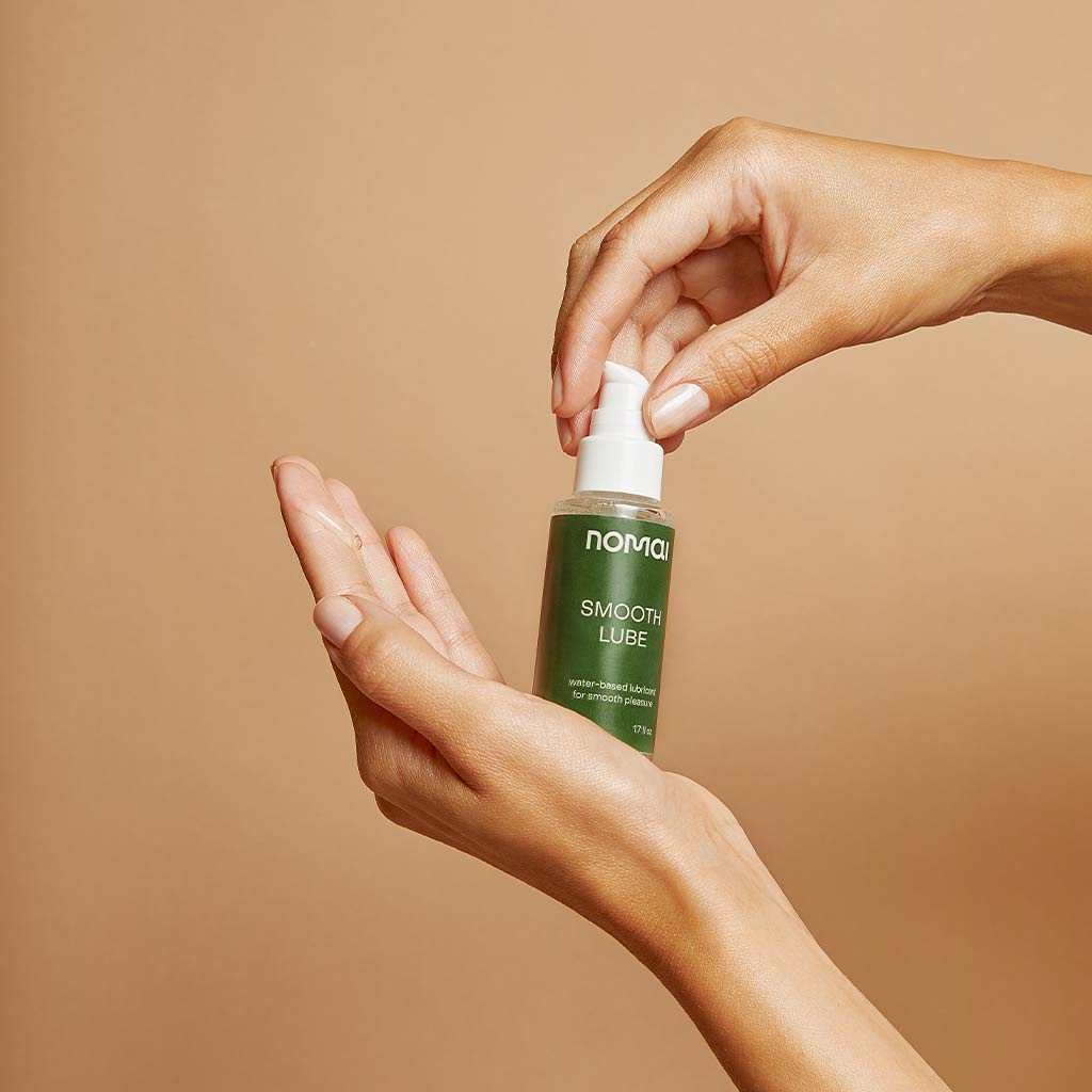 Nomai Vesipohjainen Liukuvoide; käsi pitelee vihreää muovipulloa pumppuannostelijalla, jossa lukee selkeällä groteskilla "Nomai Smooth Lube"; toinen käsi painaa pumppua, jolloin läpikuultavaa geeliä valuu avoimelle kämmenelle; pullon pinta on kiiltävä; tausta yksivärinen beige; lähikuva keskittyy käsiin ja tuotteeseen.