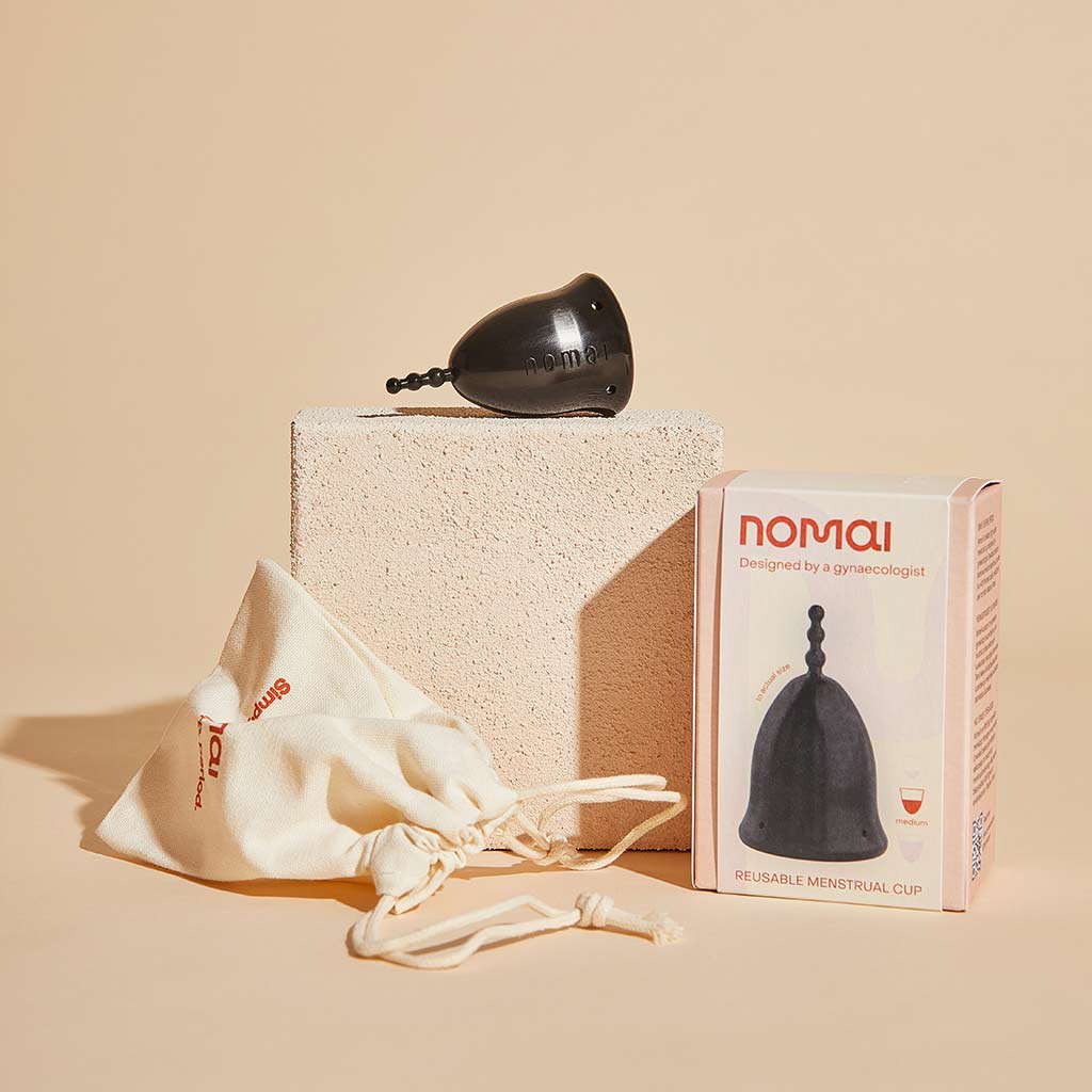 Nomai kuukuppi musta; musta, mattapintainen kuukuppi, vaaleasta kankaasta tehty vetonarupussi sekä suorakulmainen pakkaus tekstillä "Nomai Reusable Menstrual Cup"; tasainen, beige alusta ja tekstuurinen taustakuvio.