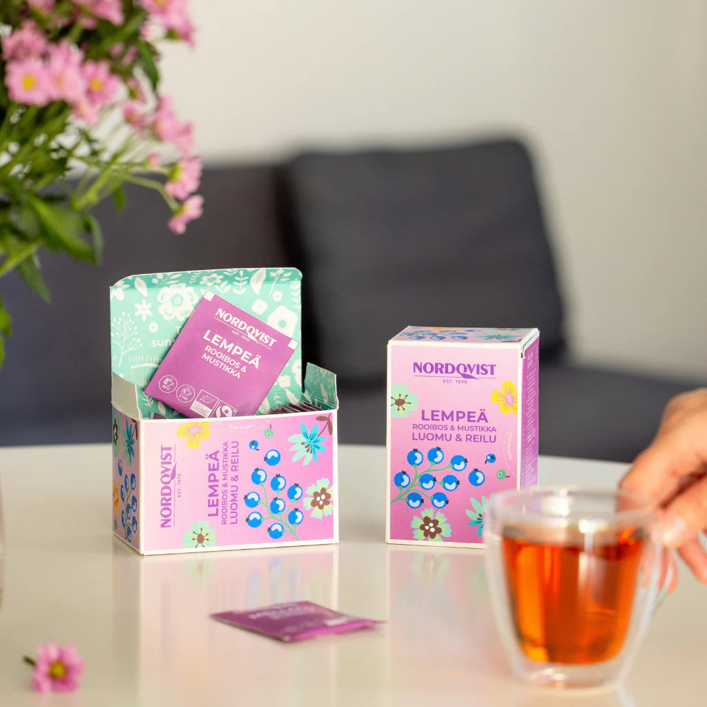 Nordqvist Lempeä Luomu Rooibos & Mustikka; käsi pitelee läpinäkyvää lasista teekuppia suorakulmaisen vaaleanpunaisen Nordqvist LEMPEÄ -teerasian lähellä, jossa näkyvissä teepusseja; yksi pussi on aseteltu lattaan pöydälle; rasia mattapinnalla, groteskikirjasin ja sana ”LEMPEÄ” selkeästi luettavissa; asetelmassa kukkia ja tarkoituksellisesti sumea tausta.