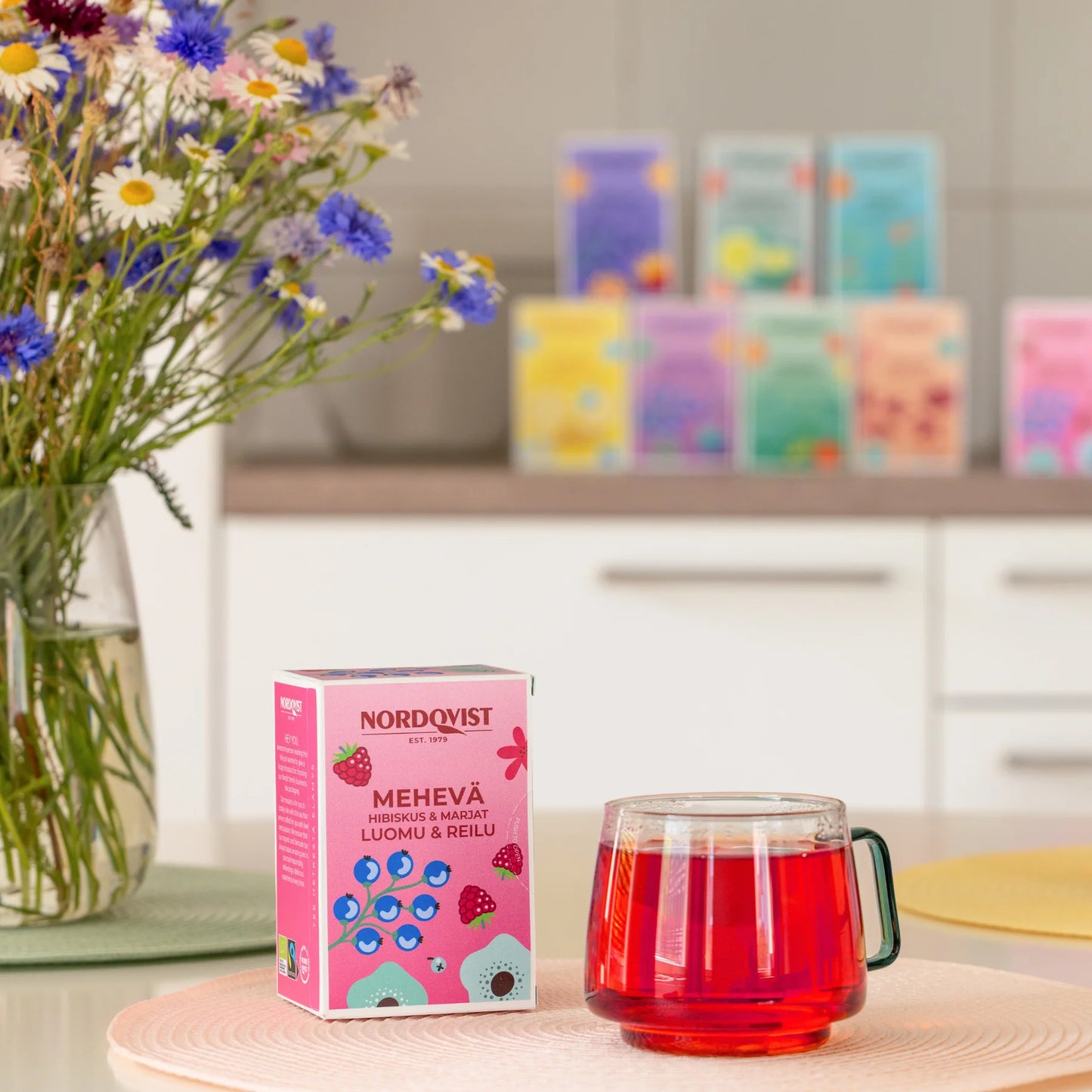 Nordqvist Mehevä Hibiskus & Marjat Luomutee: sylinterimäinen kirkas lasimuki punaista teetä ja suorakulmainen Nordqvist Mehevä -teerasia painetulla etiketillä ovat aseteltuna sileälle pinkille matolle; epätarkassa taustassa useita värikkäitä Nordqvist-teerasioita lihavoidulla kirjoituksella ja pieni maljakko sekakukkia keittiön tasolla; ei ihmisiä tai käsiä näkyvissä.