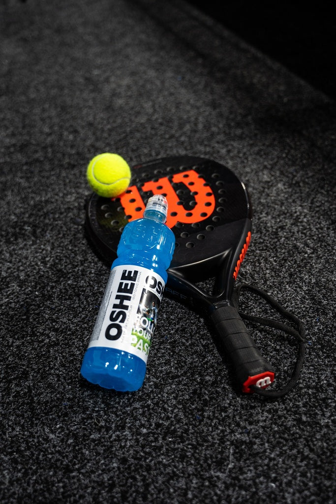 OSHEE Isotonic Multifruit 6-pack; musta-oranssi padel-maila mattapinnalla, rei'itetty lyöntipinta ja pyöristetyt reunat, lepää kirkkaankeltaisen tennispallon vieressä; sininen muovinen OSHEE-urheilujuomapullo lihavoiduin valkoisin kirjaimin seisoo pystyssä; kaikki esineet harmaalla lyhytnukkaisella matolla; ei ihmisiä tai käsiä näkyvissä; padel-maila, tennispallo ja OSHEE-pullo aseteltuna valmiina intensiiviseen peliin.