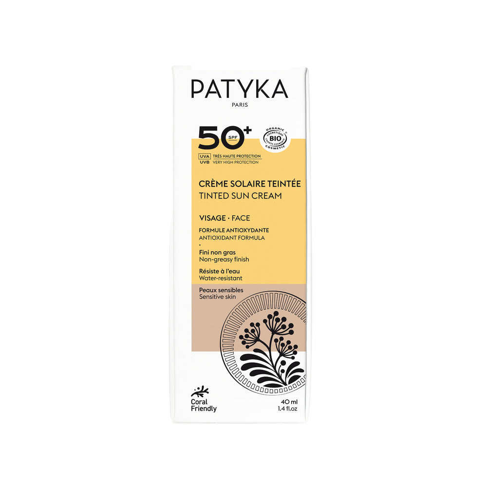 Patyka Tinted Face Sun Cream SPF50+; valkoinen laatikko Patyka-sävytettyä aurinkovoidetta kasvoille SPF 50+, mustin ja kultaisin yksityiskohdin, tekstitietoja ja kukkakuvitus etupuolella; pinta on mattapintainen; pakkaus on suorakulmainen terävin reunoin; typografia yhdistää lihavaa groteskia ja ohutta antiikvaa; etiketti mainitsee koralleja säästävän ja herkälle iholle sopivan; yksivärinen tausta, ei ihmisiä tai käsiä näkyvissä.