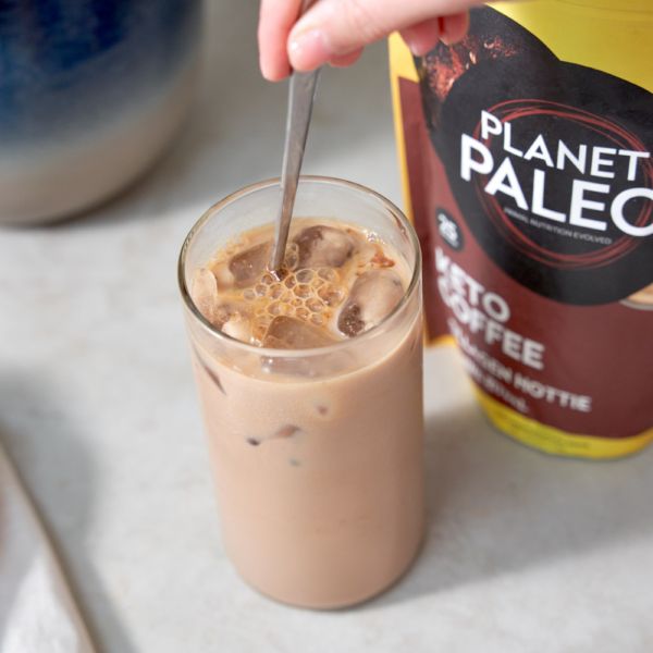 Planet Paleo Keto Coffee; käsi sekoittaa jääkahvia lasissa lusikalla, vieressä keltainen-ruskea Planet Paleo Keto Coffee -pakkaus vaalealla alustalla; lasi kirkas ja lieriömäinen, jossa näkyy jääpaloja; pakkaus mattapintainen ja suorakulmainen, lohkomainen valkoinen teksti brändinimelle, pienempi teksti alla osittain luettavissa.