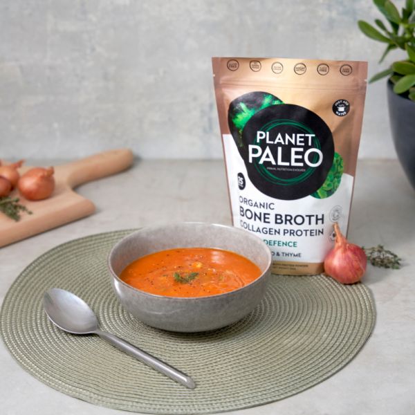 Planet Paleo Luomu Luuliemi Herbal Defence; kulho tomaattikeittoa yrtein vihreällä alustalla lusikan vieressä; taustalla mattamusta muovipakkaus tekstillä "Planet Paleo Organic Bone Broth collagen protein" lihavoiduin valkoisin ja kultaisin suuraakkosin; salottisipulia ja tuoreita yrttejä aseteltuna pienelle vaalealle puiselle leikkuulaudalle taustalla; ei ihmisiä tai käsiä; kuva otettu ylhäältäpäin.