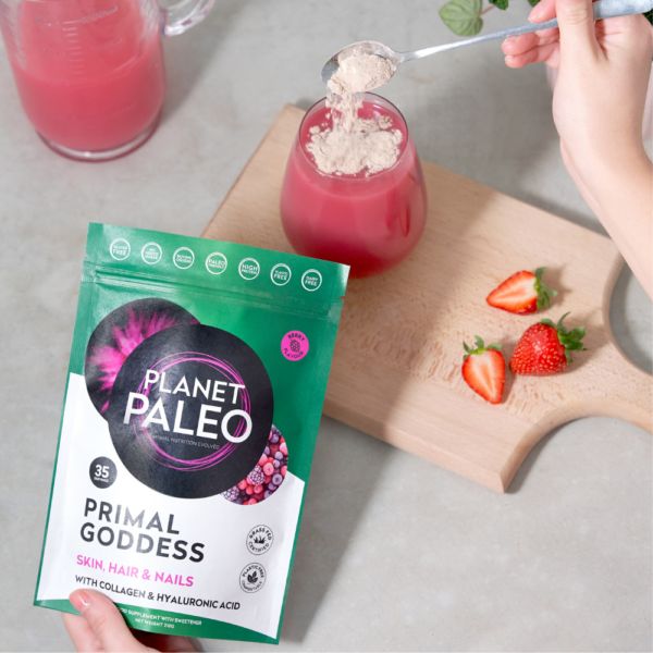 Planet Paleo Primal Goddess; henkilö lisää jauhemaista ravintolisää pussista vaaleanpunaiseen kollageenijuomaan; tuoreita mansikoita ja lasillinen eloisan vaaleanpunaista nestettä puisella tarjottimella vieressä; pussi suorakulmainen, mattapintaista joustavaa materiaalia, selkeä lihavoitu groteskiteksti; tausta neutraali ja epätarkka, viittaa sisätiloihin.