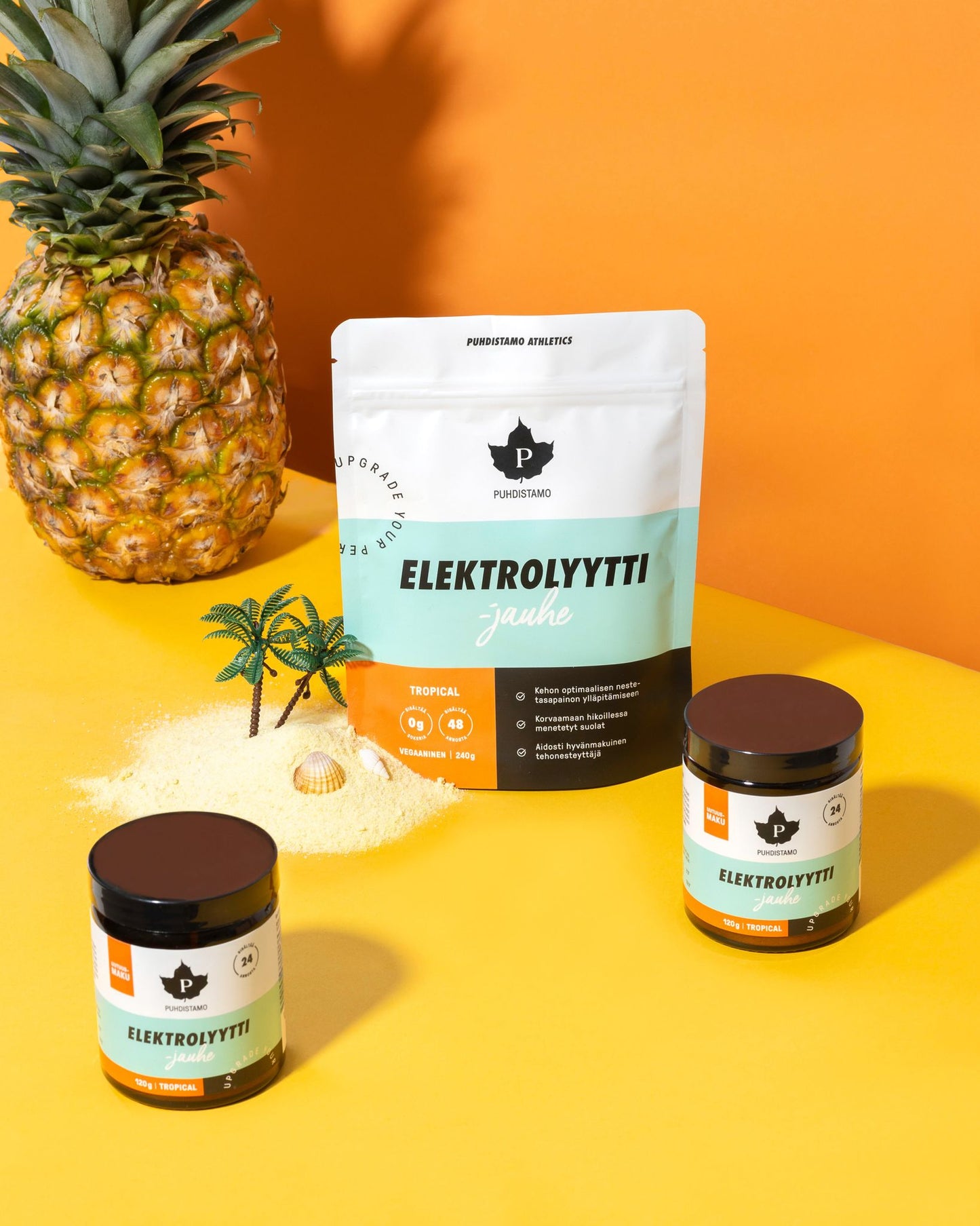 Tropical-themed display featuring a pineapple, two jars, and a large packet labeled ELEKTROLYYTTI jauhe, surrounded by sand, a mini palm tree, and a seashell on a vibrant yellow-orange background. The packet is rectangular with bold sans-serif text and matte finish. The jars are clear glass with metallic lids. All items are arranged on loose sand for a staged product shot.