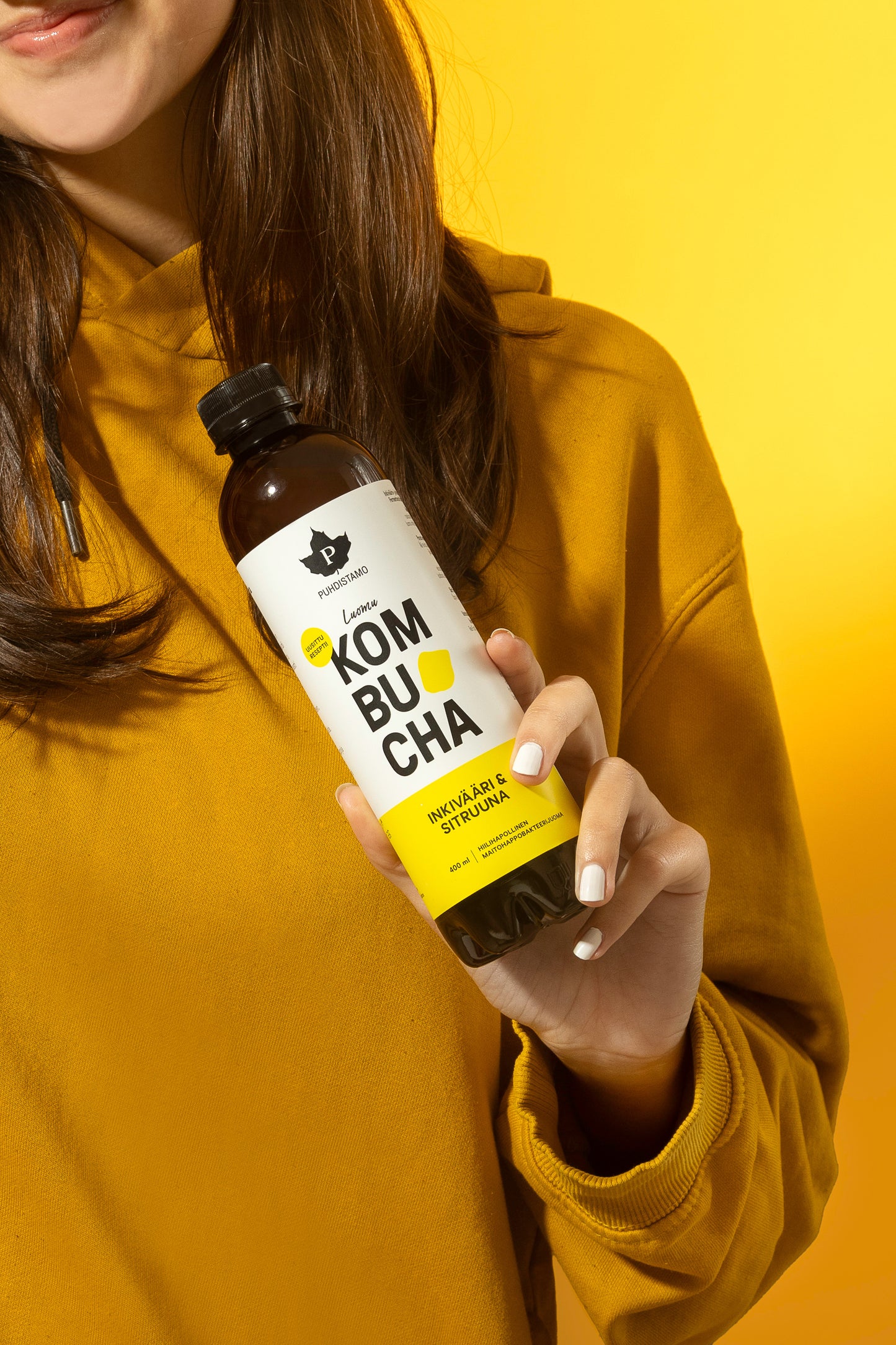 Puhdistamo Luomu Kombucha Inkivääri-Sitruuna; keltaiseen collegepaitaan pukeutunut nainen pitelee kirkasta lasipulloa, jossa inkivääri-sitruunakombuchaa; pullo sylinterimäinen, etiketissä keltaista ja valkoista väriä sekä lihavoitua, modernia typografiaa ja tuotenimi luettavissa; etiketin pinta mattaa; tausta yhtenäisen kirkkaan keltainen, tasaisesti valaistu.