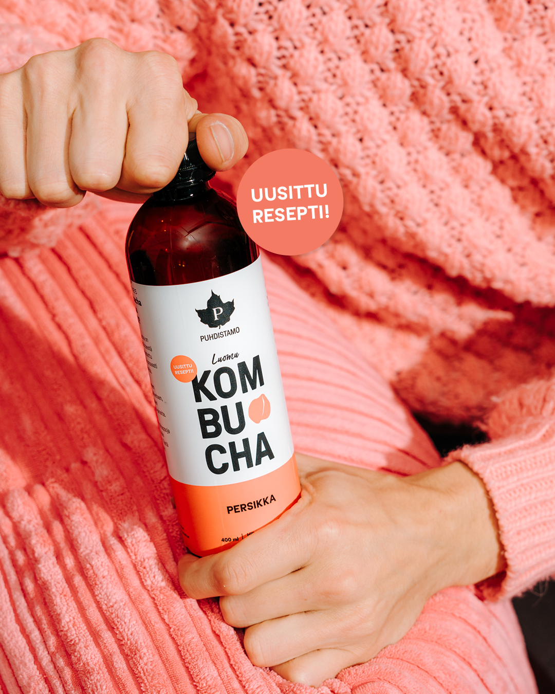 Puhdistamo Luomu Kombucha Persikka; vaaleanpunaiseen neuletakkiin pukeutunut henkilö pitelee kirkasta lasipulloa persikankombuchaa; etiketti mattapintainen, näkyvissä tuotenimi, brändilogo sekä lihavoitu teksti "UUSITTU RESEPTI!!"; tausta yhtenäisen kirkkaan vaaleanpunainen; kuvassa näkyy vain käsi ja osa vartaloa.