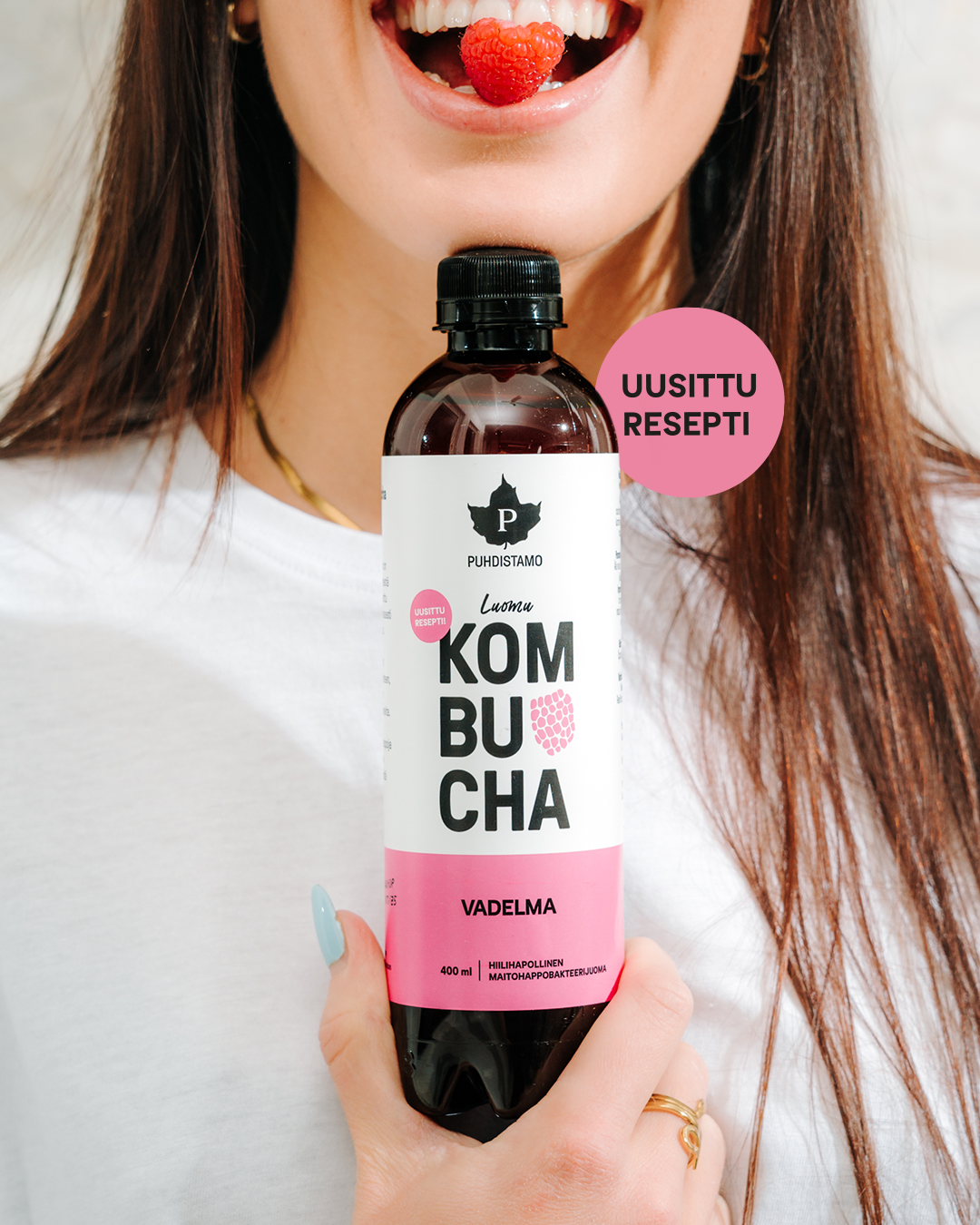 Puhdistamo Luomu Kombucha Vadelma; nainen pitelee kirkasta lasipulloa, jossa vaaleanpunaista vadelmakombuchaa; etiketissä suuraakkosin groteskitekstillä "KOMBUCHA VADELMA" sekä vaaleanpunainen ympyrä tekstillä "UUSITTU RESEPTI"; etiketti mattapintaista paperia; nainen hymyilee vadelma hampaidensa välissä; tausta neutraali ja siisti.