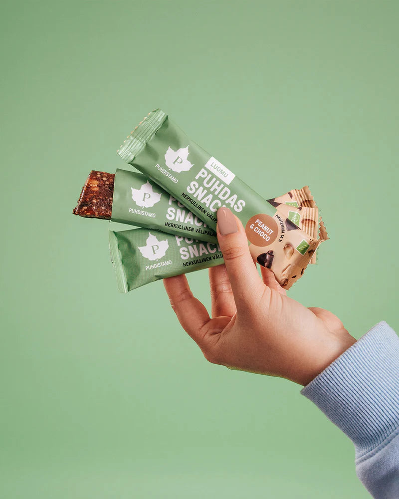 Puhdistamo Luomu Snack Peanut & Choco; käsi pitelee kolmea vihreä-beigekääreistä snackpatukkaa tasaista vihreää taustaa vasten; henkilöllä vaaleansininen pitkähihainen paita; kukin patukka suorakaiteen muotoinen ja erikseen kääritty mattapintaiseen pakkaukseen, jossa selkeä raja vihreän ja beigen osan välillä; osittain näkyvissä tuotenimiä lihavoidulla groteskikirjasimella kunkin kääreen päällä; kuva läheltä, keskittyen käteen ja patukoihin ilman muita rekvisiittoja.