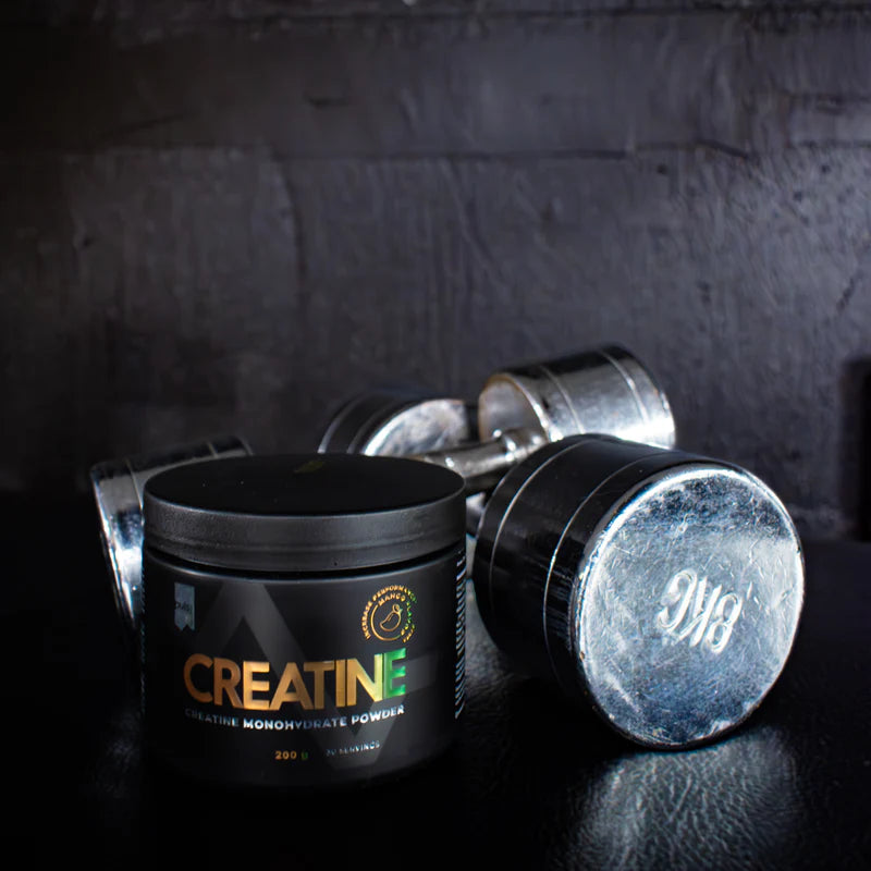 Puls Creatine, maku Mango; musta kreatiinijauhepurkki tumman pinnan päällä kahden kiiltävän metallisen käsipainon vieressä, taustalla musta tekstuuripinta; purkki lieriömäinen mattapinnalla, etiketissä valkoista lohkokirjaintekstiä, ei täysin luettavissa tästä kuvakulmasta; ei ihmisiä tai käsiä, asetelma kuvattu tasokuvana tai matalasta kulmasta.