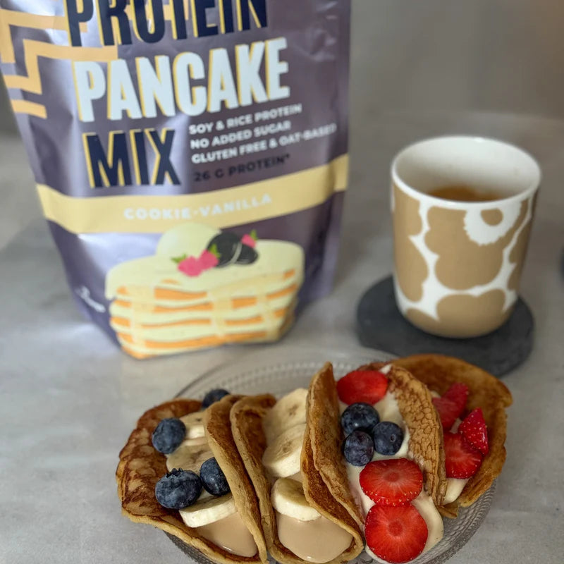 Kolme taiteltua pannukakkua täytettynä banaaniviipaleilla, jogurtilla, mustikoilla ja mansikoilla lautasella, vieressä kahvimuki. Taustalla on Cookie-Vanilla-proteiinipannukakkujauhepussi. Pussi seisoo pystyssä, mattapintainen, beigen- ja ruskeansävyinen, ja siinä on lohkomaista pääteviivatonta makutekstiä. Ihmisiä tai käsiä ei näy kuvassa. Tausta on sisätilan pöytäasetelma pehmeässä luonnonvalossa.