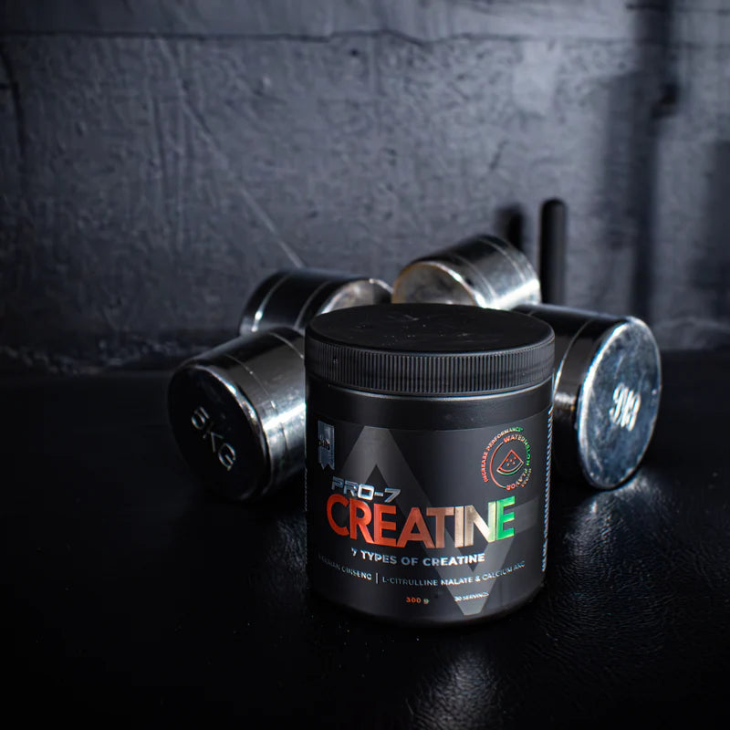 Puls Pro-7 Creatine Watermelon; musta purkki tekstillä ”Pro-7 Creatine” mustalla matalla pinnalla; sylinterin muotoisessa purkissa muovinen kierrekansi, etiketissä lihavoitu valkoinen ja punainen groteskiteksti; taustalla osittain näkyvissä kolme metallista häntahälyistä härmistä pintaa olevaa käsipainoa; hämyinen valaistus, tumma seinä taustalla, ei ihmisiä tai käsiä kuvassa.