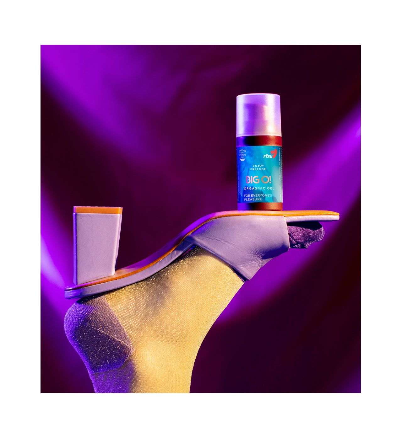A foot in a gold sock and lavender high-heeled sandal is raised, elegantly balancing a bottle of nourishing hair oil on the sole. The transparent cylindrical bottle has a white label with black text and a gold cap. No hands or other people are present. The background is purple with dramatic lighting.