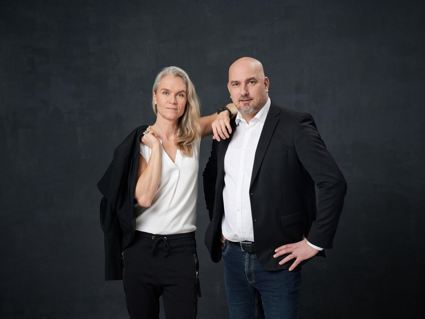 A woman with long gray hair and a man stand together against a dark background. Both wear business casual outfits; the woman has a light-colored jacket over her shoulder, while the man rests one hand on his hip. No visible product packaging, label text, or typography. Medium shot shows figures from thigh up, neutral expressions. Background is solid and untextured.