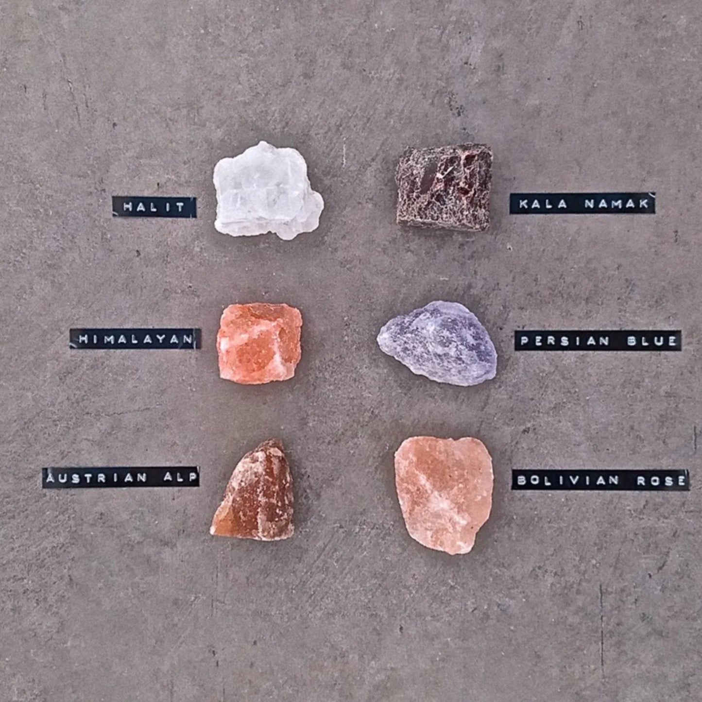 Rivsalt Suolalajitelma; kuusi eri suolakiveä, kukin merkitty: Halit (valkoinen), Kala Namak (ruskea), Himalayan (vaaleanpunainen), Persian Blue (sininen), Austrian Alp (punaruskea) ja Bolivian Rose (vaaleanpunainen), aseteltuna harmaalle pinnalle; karkeita, epäsäännöllisiä muotoja mattapinnoin; yksinkertaiset painetut etiketit näyttävät suolojen nimet; ei pakkausta tai ihmisiä näkyvissä; tasokuva ylhäältä; kokoelma esittelee maailman herkkusuolojen eloisan monimuotoisuuden.