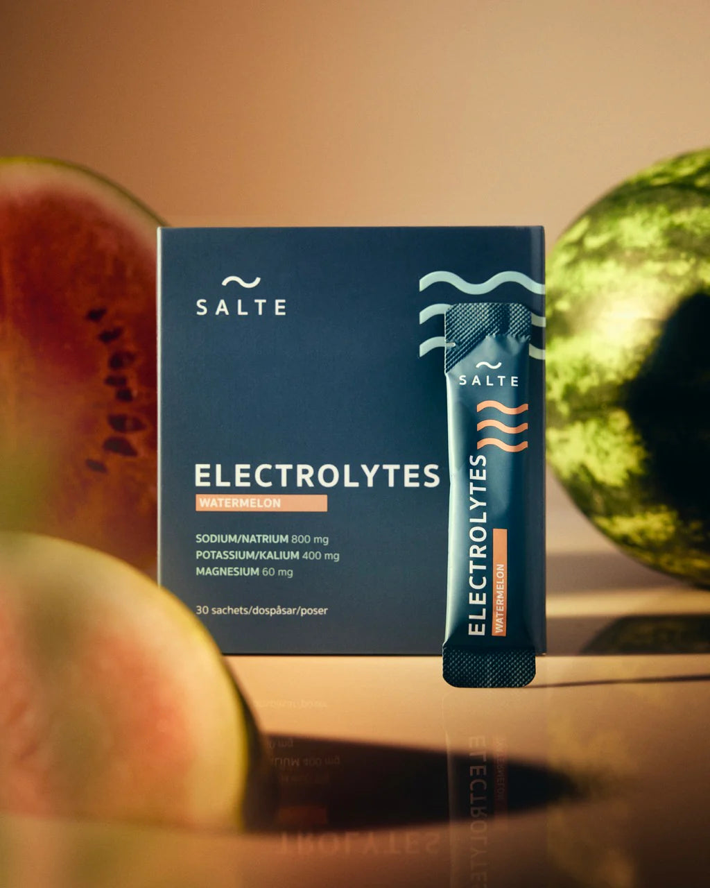 SALTE Electrolytes Watermelon -laatikko, jota vasten nojaa yksi pussukka. Pakkaus on suorakulmainen ja mattapintainen; sekä laatikossa että pussissa hallitsevat vihreä ja pinkki väri. Lihavoitu pääteviivaton typografia näyttää tuotenimen. Vesimelonin viipaleita on aseteltu tuotteiden ympärille. Tausta on pehmeästi sumennettu, mikä korostaa tuotteita etualalla. Ihmisiä tai käsiä ei näy.