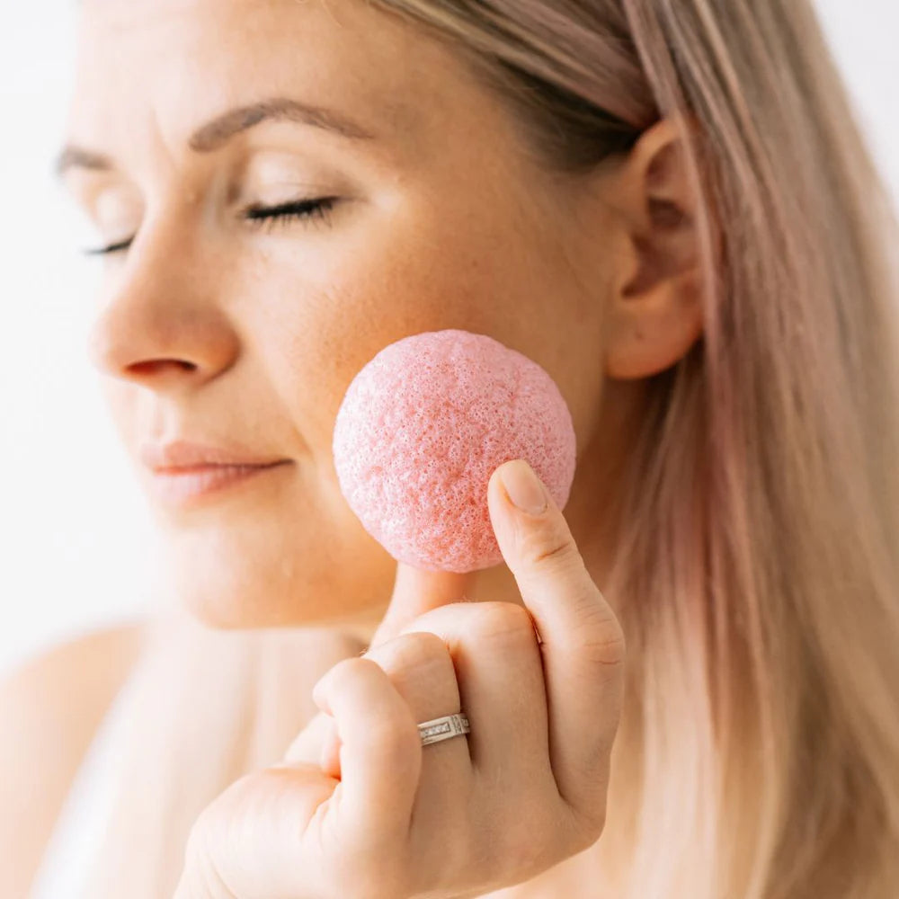 Saaren Taika Ecolution Ruusu Konjac Puhdistussieni; nainen silmät kiinni painaa hellästi pyöreää, vaaleanpunaista konjac-sientä poskeaan vasten näyttäen rentoutuneelta ja tyyneltä; sieni pyöreä, vaaleanpunainen, mattapintainen ja avattu ilman näkyvää brändäystä tai pakkausta; tausta tasainen ja tasaisesti valaistu neutraalein sävyin; kuvassa näkyy vain naisen kasvot ja käsi, iho kirkas ja tummat hiukset taaksesidottuina.