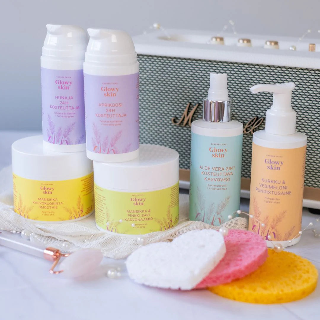 Saaren Taika Glowy Skin Mansikka Kasvokuorinta Smoothie; valikoima Glowy Skin -ihonhoitotuotteita – voiteita, emulsioita, suihkeita – sylinterin ja tuubin muotoisissa astioissa valkoisella tai pastellivärisellä pakkauksella, aseteltuna pastellisävyisten meikkisienien, metallikantisen meikkisiveltimen ja helminauhan kanssa; esineet vihreä-valkoisen Marshall-kaiuttimen edessä lihavoiduin lohkokirjaimin; ei ihmisiä eikä käsiä näkyvissä.