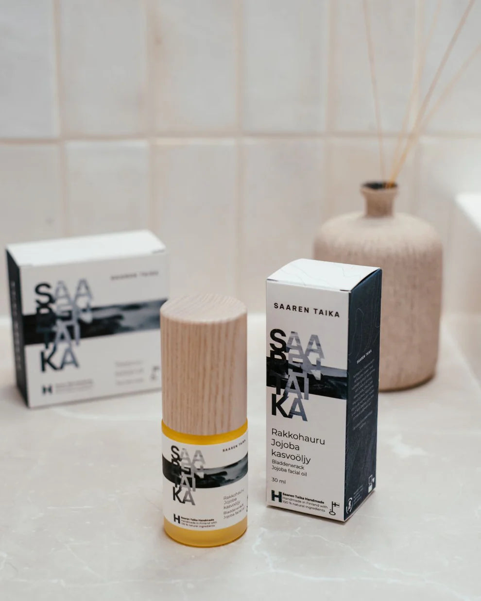 Saaren Taika Rakkohauru Jojoba kasvoöljy; pullo Saaren Taika -kasvoöljyä puisella korkilla seisoo kylpyhuoneen tasolla suorakulmaisen valkoisen laatikkonsa vieressä; pullo on kirkasta, sylinterinmuotoista lasia, sisältää vaaleansävyistä nestettä; sekä laatikossa että etiketissä lukee ”Saaren Taika” yksinkertaisella groteskifontilla; taustalla toinen samanlainen tuotelaatikko sekä beige mattapintainen maljakko, jossa hajusteosia; ei ihmisiä tai toimintaa näkyvissä.