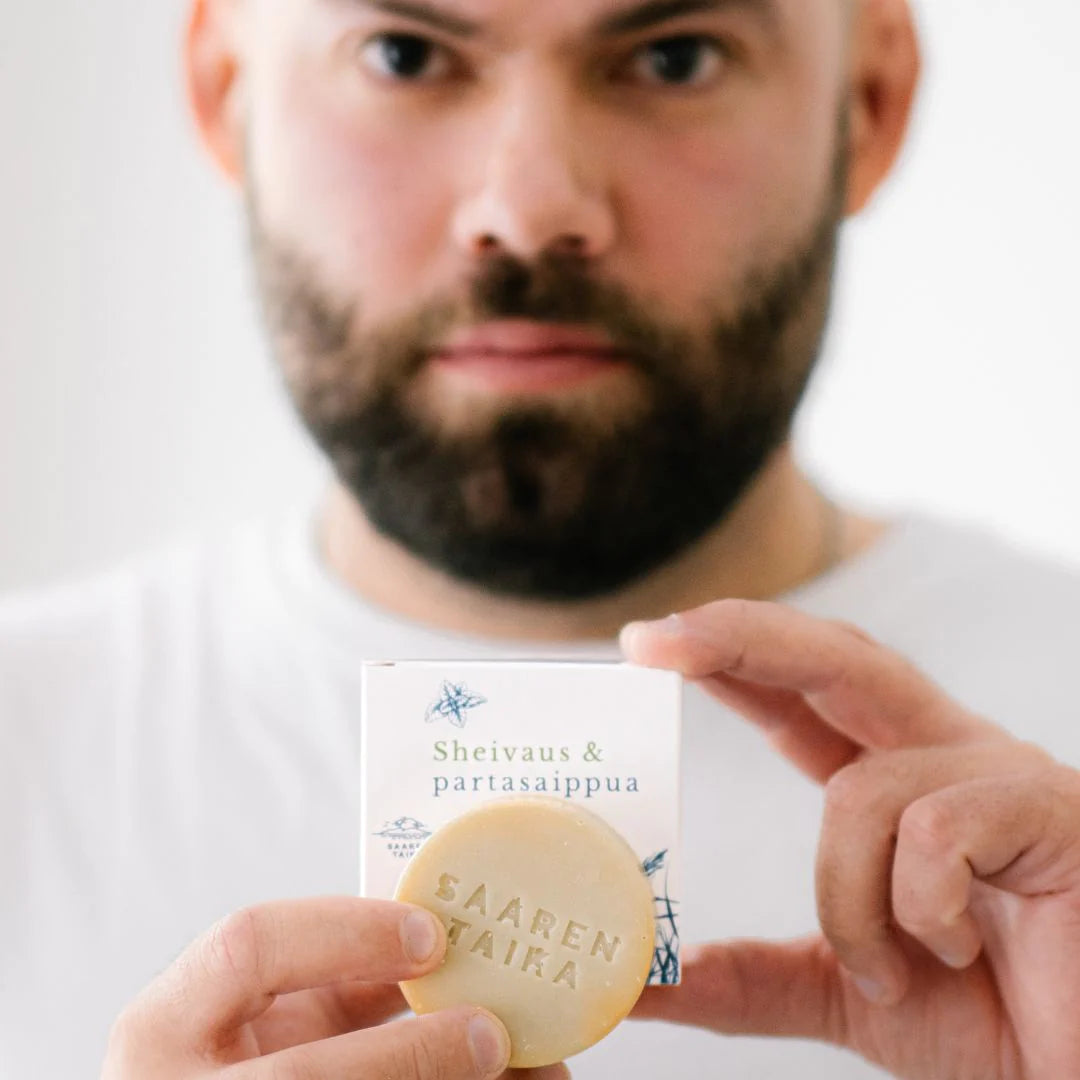 Saaren Taika Sheivaus & Partasaippua; parrakas mies valkoisessa paidassa pitelee pyöreää murrettuavalkoista saippuapalaa ja suorakulmaista laatikkoa lähellä kameraa; laatikossa painettua tekstiä "Sheivaus & partasaippua" ja "SAAREN TAIA" selkeällä sans-serif-fontilla; molemmat pinnat mattaisia; kasvot hieman epätarkat, kädet esittelevät tuotteita selvästi edessä.