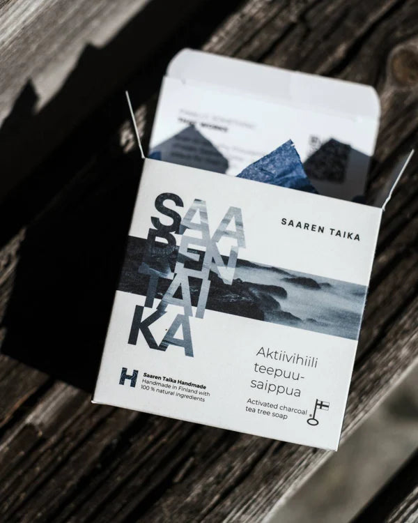 Saaren Taika Aktiivihiili Teepuusaippua; valkoinen suorakulmainen pahvipakkaus tekstillä "Saaren Taika", sisällä tumma, kiinteä aktiivihiili-teepuusaippuapala mattapintaisena; pakkaus avattuna näyttäen saippuan sisältä; selkeä groteskityyppografia; pakkaus puisella, syykuvioisella alustalla, auringonvalo ja varjo lankeavat vinosti; ei ihmisiä tai käsiä.