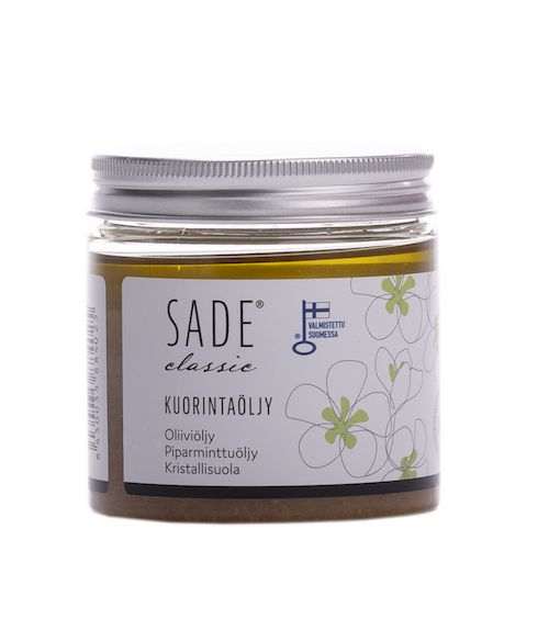 A jar of Sade Classic Kuorintaöljy exfoliating oil with a reflective silver lid, cylindrical shape, and transparent body showing pale yellow oil. The product label is in Finnish with dark green floral graphics on a white background, sans-serif fonts, and lists olive oil, peppermint oil, crystal salt. No people are present; neutral background with soft lighting.