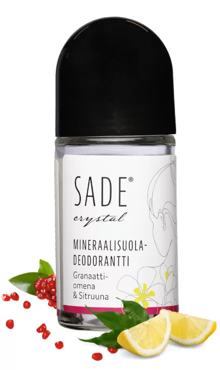 Sade Mineraalisuoladeodorantti Roll-On Granaattiomena-Sitruuna; SADE crystal -mineraalisuoladeodoranttipullo aseteltuna granaattiomenan siementen, sitruunaviipaleiden ja valkoisen kukan kera juurellaan; pullo kirkas ja lieriön muotoinen mattavalkoisella korkilla, etiketissä "SADE" lihavoidulla sans-serif-fontilla; valkoinen tausta, suoraan kuvattu; ei ihmisiä tai käsiä.