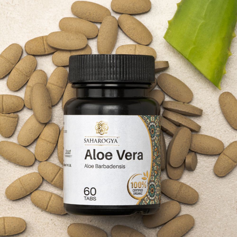 A bottle of Saharogya Aloe Vera tablets, 60 count, features a white plastic container with a green cap and matte surface, positioned upright. The label displays “Saharogya” in bold sans-serif lettering and “Aloe Vera” clearly below. White round tablets are scattered nearby along with a green aloe leaf segment on a pale textured background in a flat lay composition.