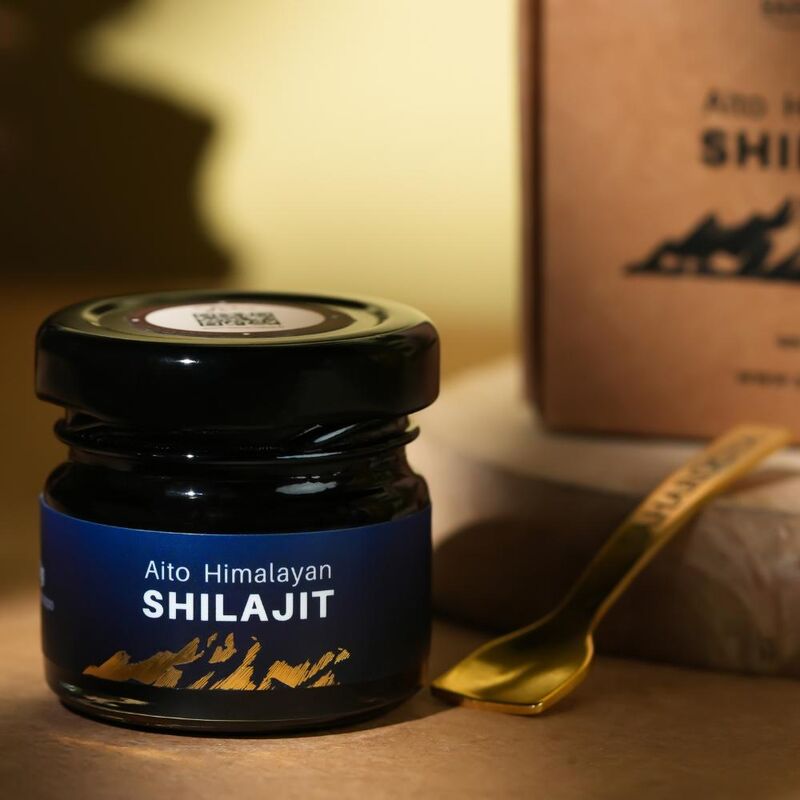 Saharogya Himalajan Shilajit pihka; pieni musta purkki tekstillä ”Aito Himalayan Shilajit” kultaisen lusikan ja vastaavan brändi-ilmeen ruskean suorakaiteen muotoisen rasian vieressä; molempien musta etiketti valkoisella groteskitekstillä; kaikki esineet vaalealla tasaisella alustalla pehmeässä valaistuksessa; ei ihmisiä tai käsiä kuvassa.