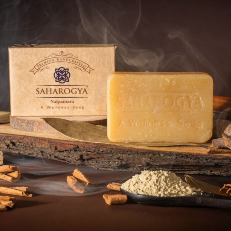 A handcrafted Saharogya cinnamon soap bar is displayed with its box, surrounded by cinnamon sticks and powder on a rustic wooden surface with soft smoke swirling in the background. The soap bar is rectangular with a matte finish in light brown color. Packaging box has minimal design and legible “Saharogya” branding in simple sans-serif font. There are no people or hands visible. The shot is taken from above.